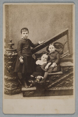 A black-and-white studio photograph of three White children on a decorative staircase: one standing on the bottom step and two seated and leaning on each other.