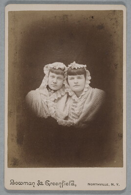 A black-and-white studio portrait photograph of two White women leaning their heads together, both wearing white shawls over their heads and shoulders.