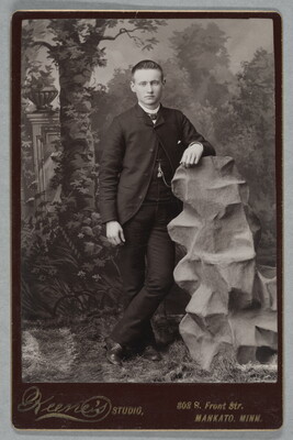 A black-and-white studio photograph of a young White man in a suit leaning with one arm on a tall, sculpted rock structure in front of a forest backdrop.