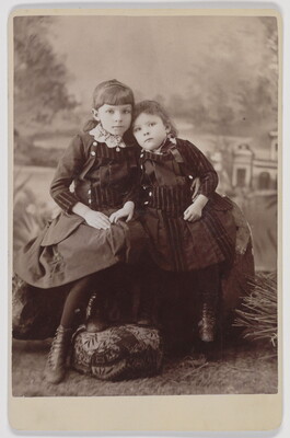 A black-and-white studio portrait photograph of two White girls seated in front of an outdoor backdrop, both wearing dark knee-length dresses.