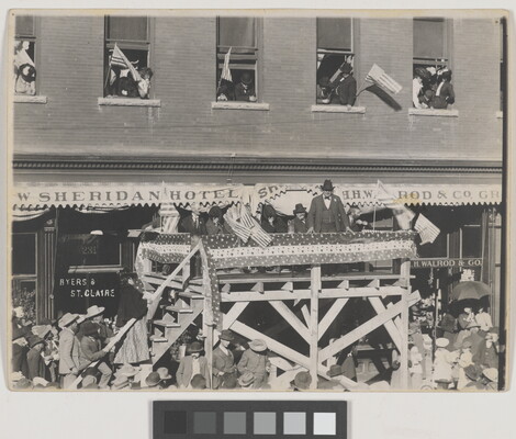 A black-and-white photograph of a group of people standing on an elevated stage in front of a building, addressing a crowd of people below.