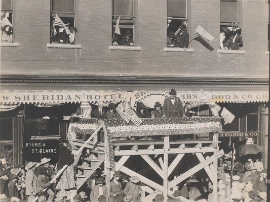 A black-and-white photograph of a group of people standing on an elevated stage in front of a building, addressing a crowd of people below.