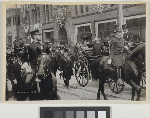 A black-and-white photograph of a parade of people on horseback, with a White man in the center in a carriage tipping his hat to onlookers.