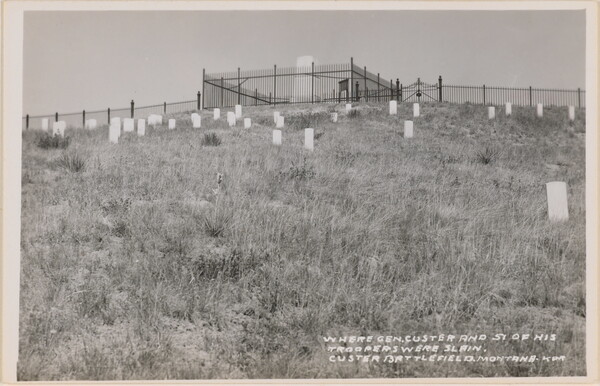 A black-and-white photograph postcard of a graveyard on a hillside below a large central monument with a fence around it.
