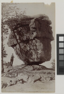A black-and-white photograph of a person standing next to a large rock formation that is balancing on a narrow end.