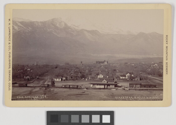 A black-and-white photograph of a railroad and station in the foreground, then a town, and snow-covered mountains in the background.