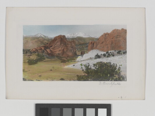A color photograph of a plain at the foot of tall, red rock formations and snowy mountains in the distance.