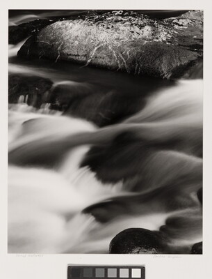 A black-and-white photograph of a water rushing over dark rocks.