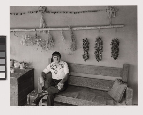 A black-and-white photograph of a man sitting on a bench under a long pole that has dried chili peppers and other herbs hanging from it.