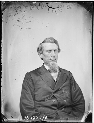 A black-and-white portrait image of a seated White man with a long goatee wearing a double-breasted jacket.