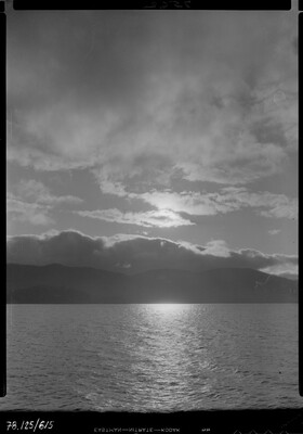 A black-and-white image of a lake, mountains on the opposite shore, under a cloudy sky at sunset.