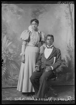 A black-and-white studio image of a seated Black man in a dark suit and a standing Black woman wearing a long-sleeved full-length, light-colored dress resting one arm on the man's shoulder.