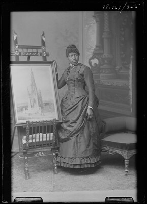 A black-and-white image of a Black woman wearing a long-sleeved Victorian-style dress standing next to an easel holding an artwork.