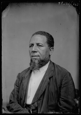 A black-and-white studio image of a seated Black man with a beard, wearing a white shirt and dark suit coat.