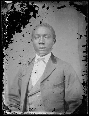 A black-and-white studio image of a young Black man wearing a three-piece suit; there is some damage to the surface of the image.