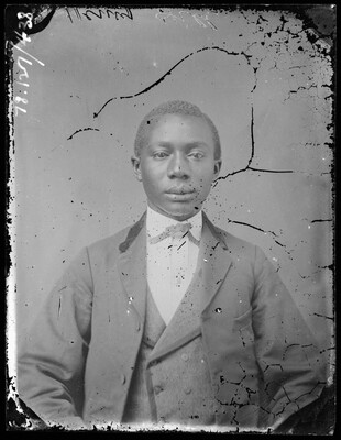 A black-and-white studio image of a young Black man wearing a three-piece suit; there are some cracks and damage to the surface of the image.