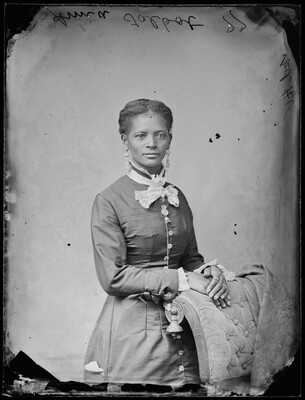 A black-and-white studio image of a standing Black woman, hair in an updo, wearing a Victorian-style dress, hands folded over a chair.