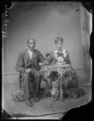 A black-and-white studio image of a Black man and woman seated by a decorative table; the man wears a suit and rests his arm on a book, and the woman wears a long dress with her hands in her lap.