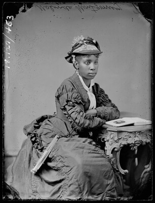 A black-and-white studio image of a seated young Black woman wearing a decorative hat and long-sleeved dress leaning on a table with her arms crossed.