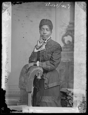 A black-and-white studio image of a Black woman, hair in an updo, wearing a long dark dress with a high collar; one hand rests on the back of a chair and the other is at her chin.