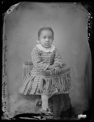 A black-and-white studio image of a toddler-aged Black girl wearing a plaid dress with a lace collar and posing on a u-shaped chair with tassels.