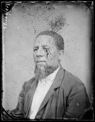 A black-and-white studio image, scratches and marks on its surface, of a seated Black man with a beard, wearing a white shirt and dark suit coat.