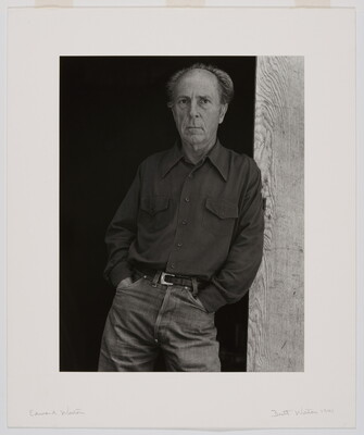 A black-and-white portrait of an older, balding White man wearing jeans and a long-sleeve button-down shirt leaning against a wooden beam.