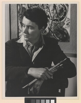 A black-and-white portrait photograph of a seated White woman holding paintbrushes in front of an abstract painting.