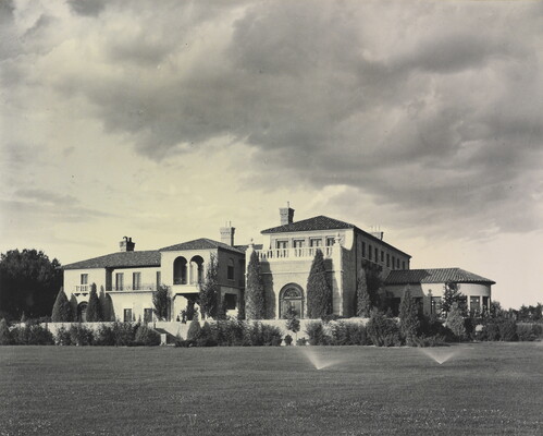 A black-and-white photograph of a large multi-story mansion with a mission-style roof and a large grass yard with sprinklers on.