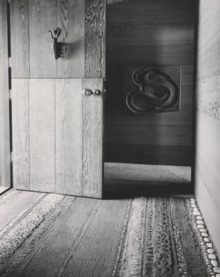 A black-and-white photograph of a Dutch door open to a decorative wood and stone floor and a carved sculpture on the far wall.