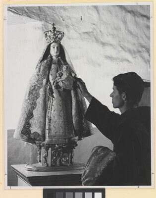 A black-and-white photograph of a friar touching a statue of the Virgin Mary dressed as queen of heaven holding the infant Jesus.