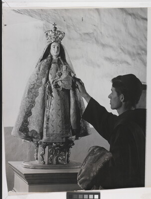 A black-and-white photograph of a friar touching a statue of the Virgin Mary dressed as queen of heaven holding the infant Jesus.