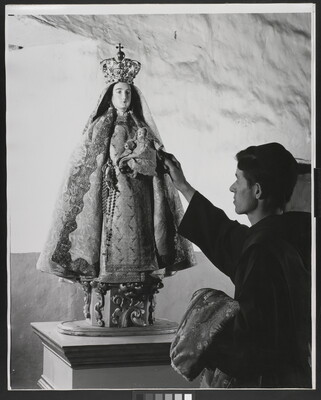 A black-and-white photograph of a friar touching a statue of the Virgin Mary dressed as queen of heaven holding the infant Jesus.