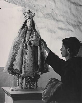 A black-and-white photograph of a friar touching a statue of the Virgin Mary dressed as queen of heaven holding the infant Jesus.