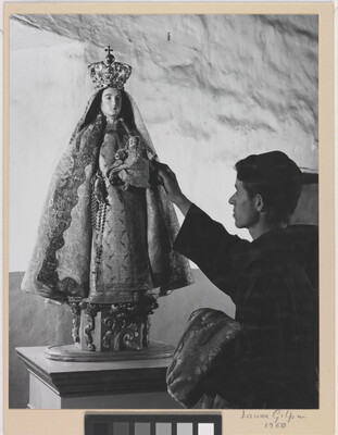 A black-and-white photograph of a friar touching a statue of the Virgin Mary dressed as queen of heaven holding the infant Jesus.