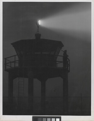 A black-and-white photograph of a person standing in silhouette atop a tower with a bright spotlight shining from the top.