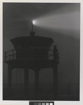 A black-and-white photograph of a person standing in silhouette atop a tower with a bright spotlight shining from the top.