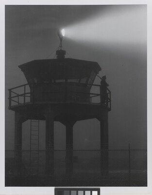 A black-and-white photograph of a person standing in silhouette atop a tower with a bright spotlight shining from the top.