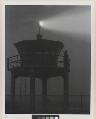 A black-and-white photograph of a person standing in silhouette atop a tower with a bright spotlight shining from the top.