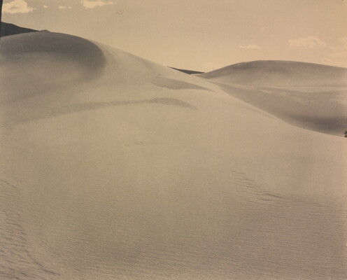 A sepia-toned photograph of a rippling sand dune rising up into a sky dotted with a few small clouds.