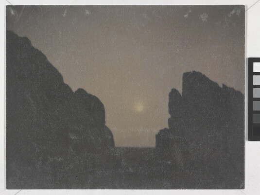 A black-and-white photograph of two rock formations in silhouette with a hazy moon in between them.