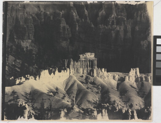 A black-and-white photograph of a landscape with hoodoos, or vertical spires of rock, on top of sand dune shapes with cliffs in the background.