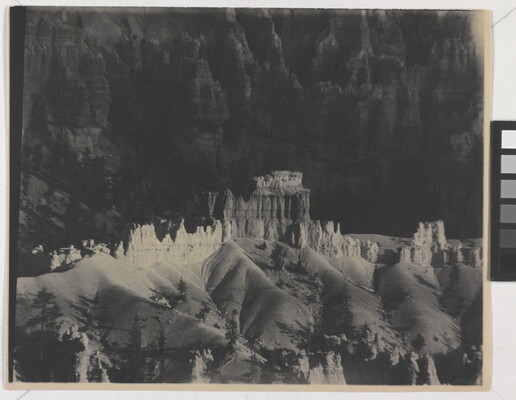 A black-and-white photograph of a landscape with hoodoos, or vertical spires of rock, on top of sand dune shapes with cliffs in the background.