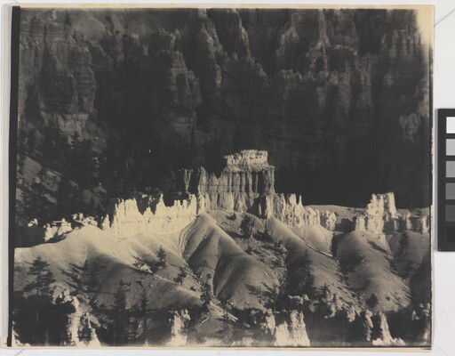 A black-and-white photograph of a landscape with hoodoos, or vertical spires of rock, on top of sand dune shapes with cliffs in the background.