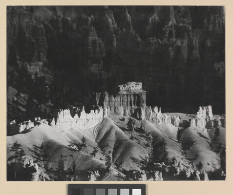 A black-and-white photograph of a landscape with hoodoos, or vertical spires of rock, on top of sand dune shapes with cliffs in the background.
