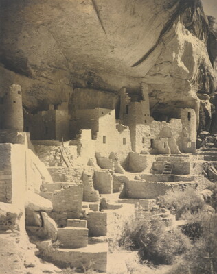 A black-and-white photograph of an abandoned, crumbling rock city built into the side of a desert cliff.