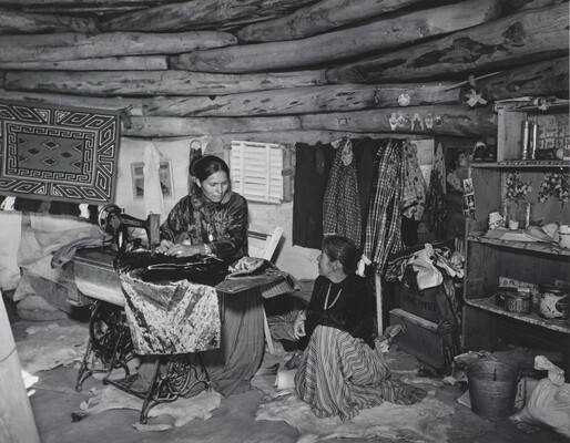 A black-and-white photograph of two Indigenous women seated, talking, and sewing inside of a low-ceilinged, wood structure.