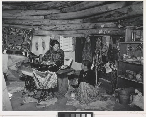 A black-and-white photograph of two Indigenous women seated, talking, and sewing inside of a low-ceilinged, wood structure.