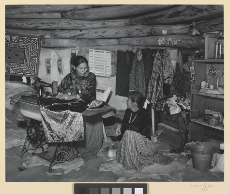 A black-and-white photograph of two Indigenous women seated, talking, and sewing inside of a low-ceilinged, wood structure.