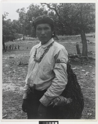 A black-and-white photograph of an Indigenous man carrying a large bag.